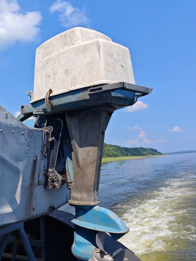 An old, weathered outboard motor is mounted on the stern of a boat, its white hood faded and cracked. The motor is attached to a blue metal bracket that connects to the boat's hull, which is also painted in faded blue and gray. The boat is moving through water, creating a white wake behind it. In the background, a tree-lined shore is visible under a bright blue sky with scattered white clouds. The scene appears to be on a body of water, likely a river or lake, given the calm water and the presence of a shoreline. The location is described as Pogodaevo, Russia, suggesting a rural or semi-rural setting. There are no people visible in the image. The overall impression is one of a functional, albeit aged, piece of marine equipment operating in a natural environment.
