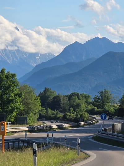 The image depicts a mountain landscape with a road network in the foreground, located in Ohlstadt, Germany. The sky is light blue with scattered white cumulus clouds, some of which obscure the peaks of the farthest mountains on the left. In the background, multiple layers of mountains rise. The most distant mountains, on the far left, are jagged, rocky, and exhibit patches of snow or ice. Closer mountain ranges are predominantly covered in dense green forest, appearing progressively bluer due to atmospheric perspective.

In the mid-ground and foreground, a system of paved roads, potentially an interchange, curves through the scene. Multiple lanes are visible, separated by white road markings, including solid and dashed lines. Silver metal guardrails line the edges of the roads, and several white and black road delineator posts are present in the immediate foreground.

An orange emergency call box, featuring an SOS logo and a telephone symbol, is mounted on a pole on the left side of the foreground, adjacent to the road. Further along the road on the right, a blue circular traffic sign with a white upward arrow indicates direction. Several unreadable dark rectangular signs are also visible on the left side of the road further back.

Lush green trees, a mix of deciduous and coniferous, frame the roads and cover the lower slopes of the mountains. In the immediate foreground, green grass and small pink wildflowers grow alongside the road. Three dark-colored passenger vehicles are visible on the roads; two are on a higher road segment, and one is on a lower, curving road section on the right, all moving away from the viewer. The scene is well-lit, suggesting either morning or late afternoon sun.