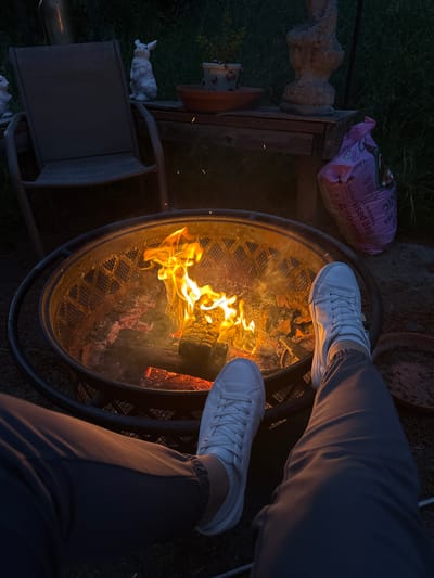 In the city of Graham, United States of America, a person sits by a fire pit at dusk. The fire pit, a circular metal structure, contains burning logs and embers, casting a warm glow on the surroundings. The person, wearing white sneakers and dark pants, is positioned with their legs extended towards the fire. To the left of the fire pit, a chair is visible, and further in the background, a wooden table holds a potted plant and decorative figurines. A bag of potting soil rests on the ground near the table. The environment appears to be an outdoor space, possibly a backyard, with grass and foliage in the background, illuminated by the fading daylight and the fire's light.