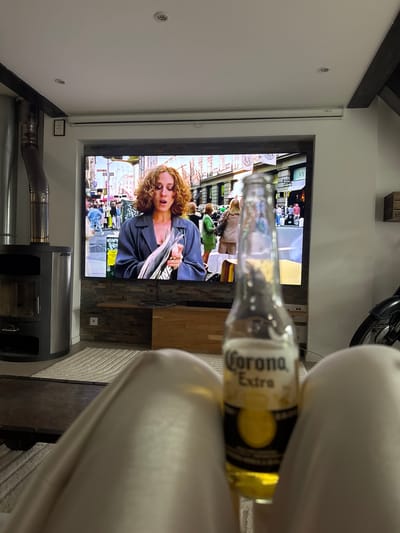 The image depicts an indoor scene with a large television screen displaying a woman with curly red hair wearing a grey blazer, holding what appears to be a newspaper or magazine. The television is mounted on a wall with stone cladding, and a wooden media console sits below it. To the left of the television, a modern wood-burning stove with a silver flue pipe is visible. A light-colored rug covers the floor in front of the television and console. In the foreground, out of focus, is a clear glass bottle of Corona Extra beer, with its label clearly visible. The perspective suggests the photo was taken from a seated position, with legs visible in the lower part of the frame. The overall impression is of a person relaxing at home, watching television and drinking a beer. There is no visible indication that this scene is located in Mandelieu-la-Napoule, France.