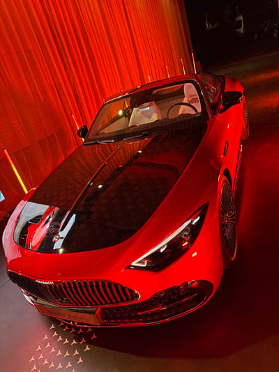 A red Mercedes-Maybach Vision 6 Cabriolet concept car is centrally displayed indoors in Herentals, Belgium. The vehicle features a vibrant red body with a contrasting glossy black hood, which subtly reveals a pattern of Maybach logos under reflections and is bisected by a chrome strip. The front fascia includes a prominent chrome vertical-slat grille with "MAYBACH" lettering above it and a "Mercedes-Maybach" plaque on the bumper. Elongated LED headlights are visible. The car is equipped with large, multi-spoke wheels. The interior, visible through the windshield, shows light-colored, possibly white or cream, quilted seats with an embossed Maybach logo. The immediate background consists of a red-illuminated pleated fabric curtain, accompanied by vertical light strips. The dark floor beneath the car features illuminated or stylized projections of the Maybach "double-M" logo pattern. No people or active interactions are visible in the scene.