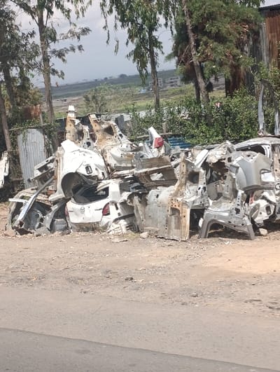 The image displays a roadside scene in Matungulu west ward, Kenya, featuring a substantial accumulation of dismantled vehicle components. In the immediate foreground, a grey paved road transitions to a light brown dirt shoulder. The central focus is a large, disorganized pile of automotive scrap, consisting primarily of stripped vehicle body shells and skeletal chassis structures. These parts are predominantly white and grey metal, exhibiting corrosion and damage from disassembly. A prominent element within the pile is the rear section of a white hatchback or SUV, identifiable by its tail lights and a partially visible 'Mazda' emblem. The background reveals a line of tall, slender trees with sparse green foliage, a corrugated metal fence on the left, and a distant valley landscape with green fields and scattered buildings, all under an overcast sky. No individuals, actions, or active processes are observed within the frame.