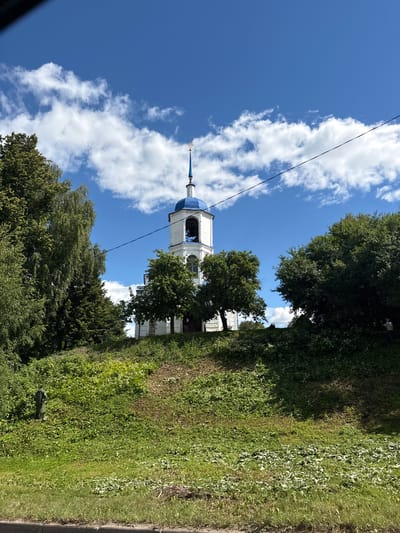 The image shows a white church with a blue domed belfry and a golden cross atop it, set against a bright blue sky with scattered white clouds. The church is situated on a grassy hill, surrounded by lush green trees. In the foreground, there is a slope covered in grass and various plants. A black electrical wire stretches across the upper part of the image, passing in front of the church. To the far right, a person in a blue shirt is visible, seemingly trimming vegetation with a tool. The overall scene appears to be a tranquil, daytime view of a religious building in a natural setting. The church depicted is likely the Church of the Transfiguration (Preobrazhenskaya Church) in Pereslavl-Zalessky, Russia, given its architectural style and the presence of a blue dome and belfry, common features in Orthodox churches of that region.