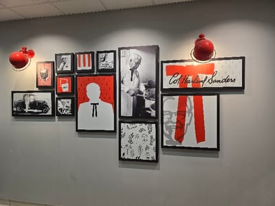 The image displays a gallery wall inside a building, likely a KFC restaurant, located in Longeville-lès-Saint-Avold, France. The wall is painted a neutral gray and adorned with a collection of framed art pieces centered around Colonel Harland Sanders and KFC branding.
Two red, spherical wall lights with metal bases illuminate the display from above. The artwork comprises various styles, predominantly using a black, white, and red color palette.
Key framed pieces include:
- A large black and white portrait of an elderly Colonel Sanders in a chef's jacket, positioned prominently towards the upper-middle right.
- A large graphic silhouette of Colonel Sanders with a bow tie, depicted in white against a red background filled with small red "T" shapes, located centrally at the bottom.
- A horizontal frame showcasing "Col. Harland Sanders" in elegant script over a red bar, set against a white background patterned with small "T" shapes, situated in the upper right.
- A large square graphic featuring a gray outline of Colonel Sanders' face on a white background, partially obscured by two bold red vertical stripes, located below the "Col. Harland Sanders" text.
- A black and white photograph of a vintage car in the bottom left.
- Various smaller frames:
- One with a red background and a white silhouette of a KFC bucket and the letters "KFC".
- Several black and white photographs, some depicting people and others with architectural or abstract details, though specific subjects are unclear due to resolution.
- A red and white graphic with vertical stripes.
- A stylized black and white pattern of leaves or foliage in the bottom-middle.
- A graphic with a chicken/rooster silhouette against a red and white background.
No specific activity or event is taking place; the image captures a decorative static display. The interior setting suggests artificial lighting, and there are no discernible details about the time of day or weather. The overall impression is a modern tribute to the KFC brand and its founder.