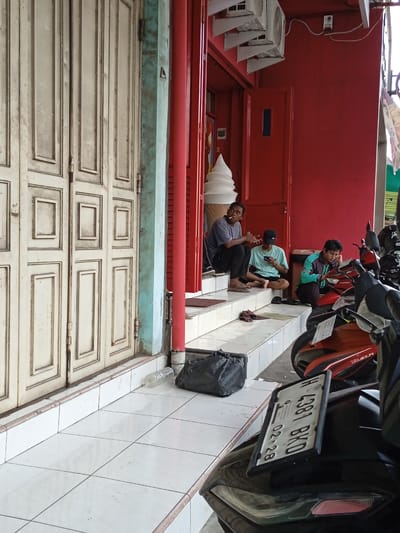 The image shows a street scene in Kauman, Indonesia. Three young men are sitting on the steps of a red-painted building, which appears to be a commercial establishment with a large, decorative ice cream cone on its facade. The man on the left is smoking a cigarette. The man in the middle is looking at a mobile phone. The man on the right is reading. Several parked motorbikes are in the foreground and to the right, with their license plates visible. The building to the left has a large, multi-paneled wooden door with a distressed, painted finish. A black plastic bag and a clear plastic bottle are near the base of the steps. Air conditioning units are installed above the building's entrance. The area has tiled flooring.