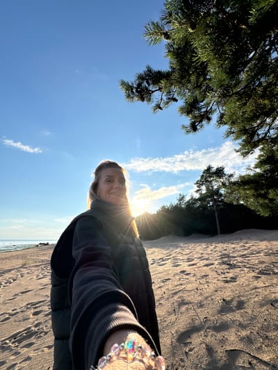 A woman stands on a sandy beach, facing the camera. She is wearing a dark puffer vest over a dark long-sleeved shirt. Her hair is blonde and pulled back. She is smiling. The sun is low in the sky, casting a warm glow and lens flare. Behind her, the sea is visible in the distance, and trees line the back of the beach. Footprints are visible in the sand. The image appears to be taken in Shepelevo, Russia, as indicated by the context.