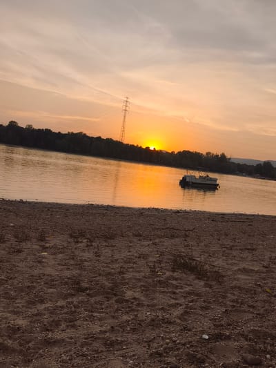 This image captures a serene sunset over a body of water, likely a river, in Dunakeszi, Hungary. The foreground is a sandy beach with scattered pebbles and sparse vegetation, suggesting a natural, undeveloped shoreline. The water itself reflects the warm, golden hues of the setting sun, creating a shimmering surface.
Dominating the mid-ground is a dark line of trees forming the opposite bank of the river, with a tall, lattice-like transmission tower visible against the vibrant sky. Further in the distance, the landscape appears hilly and forested, fading into the twilight.
A small pontoon boat is anchored in the water, adding a touch of human presence to the otherwise natural scene. Its white structure stands out against the orange and yellow reflections.
The sky is a gradient of soft oranges, yellows, and pale grays, with wispy clouds that hint at a clear evening. The sun itself is a bright disc just above the tree line, casting a warm glow over everything. The overall mood is peaceful and tranquil, evoking the beauty of nature at dusk. The time of day is clearly sunset. There is no visible text in the image.