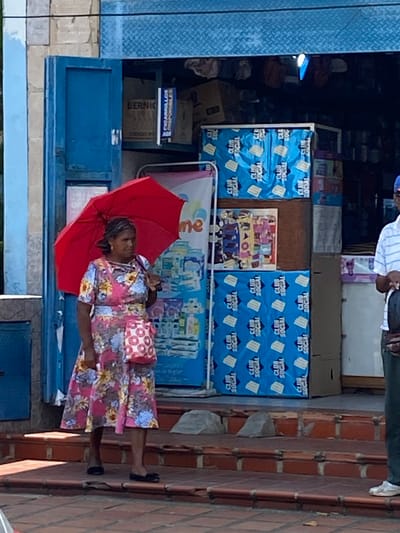 The image shows a woman standing in front of a small shop in San José de Barlovento, Venezuela. She is holding a red umbrella and wearing a floral dress with a pink patterned purse. The shop has blue walls and window. Several items are visible on shelves including boxes labeled "CLUB SOCIAL" and "BERNIC CIGARRILLOS". A person wearing a striped shirt is visible above the shop. The building is constructed with brick.