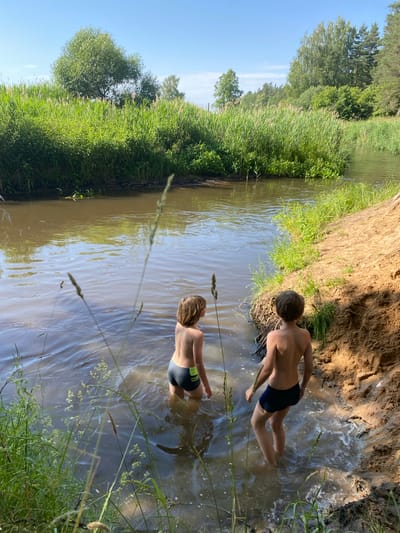 In this media file, two young boys are wading in a shallow, muddy river on a sunny day. The scene is set in a natural environment with lush green vegetation on the riverbanks, including tall grasses and trees. A sandy bank is visible on the right side of the image. The boys are wearing swim trunks, and the water is murky and reflects the surrounding greenery and sky. The sunlight suggests it is midday. The overall atmosphere appears relaxed and playful, indicative of a summer outing. There is no visible text in the image. The location context provided is Кліноцкі сельскі Савет, Belarus.