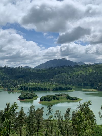 This image depicts a serene natural landscape in the vicinity of Kuda Maskeliya, Sri Lanka. A calm, jade-colored lake dominates the lower half of the frame, with two small, tree-covered islands situated in its center. The foreground features a dense collection of tall, slender trees and verdant foliage, suggesting a lush, mountainous environment. Beyond the lake, rolling hills stretch into the distance, covered in a tapestry of dense greenery, likely tea plantations and forests, interspersed with scattered trees. A distant, hazy mountain range forms the backdrop. The sky above is a dramatic mix of bright blue patches and heavy, greyish-white clouds, indicative of a dynamic weather system. There are no people or man-made structures prominently visible in the scene, emphasizing the natural beauty of the location.