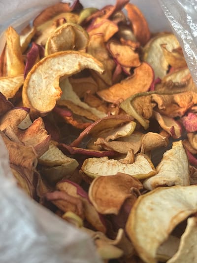 A close-up, overhead perspective captures a substantial quantity of irregularly shaped dried apple slices within a translucent plastic bag. The slices display a range of colors, from pale yellow and cream to reddish-brown and deep red, indicating the presence of apple skin on some pieces. The texture is shriveled and dry, characteristic of dehydrated fruit. The dried apple pieces are densely packed, occupying nearly the entire visual field. Edges of the clear plastic bag are visible along the upper left and right sides of the frame. No individuals, actions, or other discernible objects are present in the scene, which is located in Ivanovka, Russia.