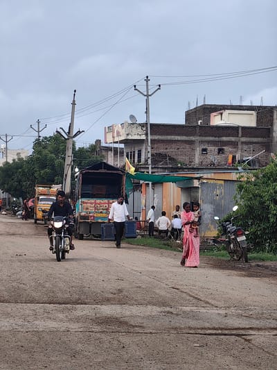 In Walsawangi, India, a dusty road stretches ahead under an overcast sky, with a line of utility poles and power lines bisecting the frame. Buildings with brick and concrete facades line the right side of the road, some appearing incomplete. A woman in a pink sari holds a young child in her arms on the right side of the road, near a parked motorcycle. On the left, a man on a motorcycle rides toward the viewer, while behind him, a yellow auto-rickshaw and a large truck are visible. Another man walks away from the truck, carrying two blue crates. Several other individuals are present in the background, some standing and others seated. Lush green trees are visible on the left side of the road.