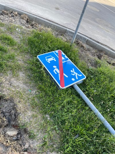 A blue traffic sign with white pictograms of a car and people playing is lying on its side in the grass next to a road in Захарьино, Russia. The sign is damaged, with a red stripe across the middle. The pole supporting the sign is also bent. The surrounding environment consists of grass, dirt, and a paved road with manhole covers. The scene appears to be an outdoor public space.