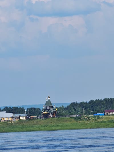 The image shows a body of water in the foreground, with ripples on the surface. In the middle ground, a green, grassy bank rises, leading up to a settlement. Several buildings are visible, including a distinctive wooden church with green roofs and golden domes, and several houses. Behind the settlement, a dense forest stretches across the horizon, with rolling hills in the far distance. The sky above is a pale blue with scattered white clouds. The scene is tranquil and appears to be a rural setting.