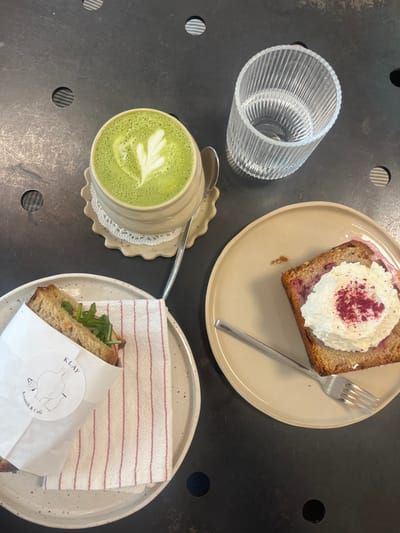 The scene depicts a tabletop arrangement of food and beverages at KLAY Keramik & Café, located at Eisenbahnstraße 19, 67655 Kaiserslautern, Germany. The dark, textured table surface features a variety of items. In the upper center, a beige ceramic mug with a ridged exterior contains a bright green matcha latte, distinguished by a white leaf-patterned latte art. The mug rests on a matching ruffled ceramic saucer, with a white doily underneath. A silver spoon is positioned on the saucer. To the upper right, a clear, vertically ribbed glass tumbler holds a clear liquid. On the lower left, a round, speckled light-colored ceramic plate presents a sandwich, partially wrapped in white paper bearing the "KLAY Keramik & Café" logo. Visible sandwich components include whole-grain bread and green leafy vegetables. A folded white napkin with thin red stripes is placed beneath the sandwich wrap. On the lower right, an irregular beige ceramic plate holds a slice of cake or loaf, exhibiting red fruit pieces within its light-brown structure. This item is topped with a substantial dollop of white cream, sprinkled with red powder. A silver fork rests diagonally across this plate. No individuals are visible in the frame.