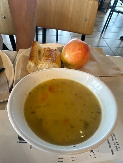 The image displays a meal served on a white tabletop within an indoor dining establishment in Medford, United States. In the immediate foreground, a white ceramic bowl contains a creamy, light yellow-green soup with visible orange (likely carrots) and green vegetable pieces. To the left of the soup, a white rectangular plate holds a torn piece of light brown, crusty bread. Adjacent to the bread on the same plate, and resting on a light brown paper napkin, is a red and yellow apple. A white paper placemat with black text is partially visible beneath the soup bowl. The background is blurred, showing parts of wooden chairs with metal frames and a light-skinned person's bare leg on the far left. The floor appears to be light-colored tile.