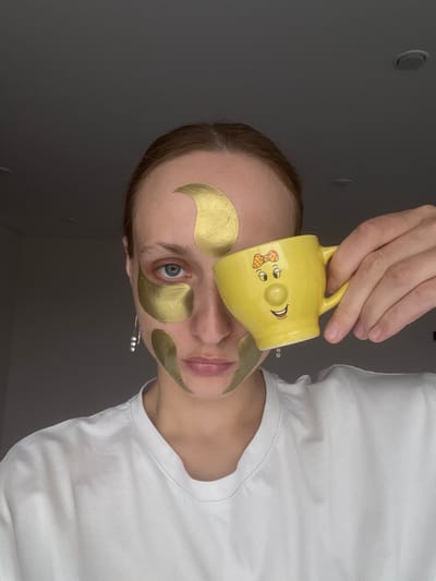 This indoor close-up image, taken in Sepych, Russia, features a person engaged in a self-care routine. The subject, likely female, has fair skin and light brown hair, and is wearing a plain white short-sleeved t-shirt. Their face is decorated with multiple golden, crescent-shaped hydrogel patches placed under both eyes, on the forehead, and near the temples.
The person holds a bright yellow ceramic mug with both hands, humorously positioning it over their upper face, obscuring part of their forehead and one eye. The mug features a cartoonish face design, including eyes (one winking), a round nose, and a wide, smiling mouth, with a small red polka-dot bow depicted near one eye.
The subject's visible right eye, which peeks out below the mug, is open and looks directly towards the viewer, revealing a blue iris. A slender earring with three small, pearl-like beads hangs from their right ear. The background is a plain, dark gray wall or surface, suggesting an indoor setting. Two faint circular indentations, possibly recessed lights or vents, are visible on the upper left side of the background. The scene conveys a casual and possibly lighthearted moment of personal pampering. No visible text is present in the image.