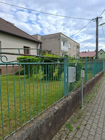 The image shows a residential street in Ilava, Slovakia, on a cloudy day. A weathered green metal fence runs along the side of a paved sidewalk. Beyond the fence, a patch of grass is visible, bordered by a concrete wall. Behind the grass and fence, a light-colored house with a brown-tiled roof is partially visible on the left. Further back, a grey building with white-framed windows and balconies stands, and in the distance, red-tiled roofs of other houses can be seen. A concrete utility pole with electrical wires is situated to the right of the grey building. The overall scene depicts a suburban or small-town environment.