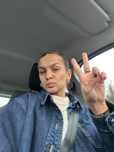 The image features a young adult female seated in the driver's seat of a vehicle, positioned centrally and facing the camera directly for a selfie. She has light skin, dark hair pulled back, and light-colored eyes. Her lips are pursed, and her left hand is raised, displaying a "peace" sign with the index and middle fingers extended. She wears a dark blue denim collared jacket over a cream-colored chunky knit sweater. Her left ear is adorned with a gold-toned, multi-pronged earring, and a similar earring is visible on her right ear. A gold-toned ring is on her left ring finger, and a gold-toned chain-link bracelet is on her left wrist. Her fingernails are painted a dark shade, possibly plum or dark grey.
The interior environment is consistent with a modern automobile. A dark grey seatbelt crosses her torso diagonally. Behind her, a dark grey headrest from the driver's seat is visible. The car's grey ceiling and a grey sun visor are visible in the upper right. Through the vehicle's windows, primarily the passenger-side window and partially the rear window, an indistinct outdoor environment can be seen, characterized by the dark shapes of trees and a cloudy, overcast sky. No specific landmarks or architectural features identifiable with a precise location within Saint-Cyr-sur-le-Rhône, France, are discernible.