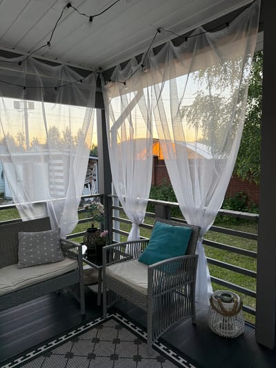 The image depicts a covered patio or porch in the city of Alyoshkino DP, Russia, during the evening golden hour. The setting appears to be a residential area, possibly a dacha (summer house) given the outdoor furniture and rustic elements.
The foreground is dominated by a dark wooden deck, partially covered by a patterned rug. On the deck are two wicker chairs. The chair on the left has a grey cushion with white stars and a light grey seat cushion. The chair on the right has a turquoise cushion and a light grey seat cushion. Between the chairs is a small dark table holding a dark vase with pink flowers. To the right of the right chair, a white woven lantern with a rope handle sits on the deck.
Behind the furniture, a metal railing encloses the porch. Beyond the railing, a grassy area is visible. The porch ceiling is white wood, from which string lights with exposed bulbs are hung. Translucent white curtains are draped from the ceiling, obscuring the view of the surroundings but allowing the warm glow of the sunset to filter through. In the background, beyond the curtains and railing, there are glimpses of buildings and trees silhouetted against the bright orange and yellow sky. The overall atmosphere is peaceful and inviting, suggesting a relaxed outdoor living space. There are no people present in the image.