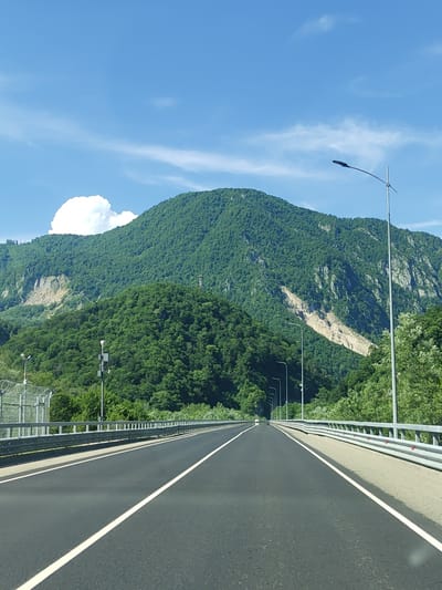 The image captures a view from a highway, likely in or near Monastyr, Russia, given the context. The road, paved in dark asphalt, is marked with white lane dividers and stretches towards a large, verdant mountain range. The mountains are densely covered with trees, with some rocky, lighter-colored sections visible on their slopes, suggesting potential geological activity or erosion. A bright blue sky with scattered white clouds forms the upper portion of the scene. Along the sides of the highway, metal guardrails are prominent. Several streetlights with dark fixtures are spaced along the roadside. To the left, a security fence with barbed wire is also visible. The overall impression is a scenic, remote highway passing through a mountainous, forested region.