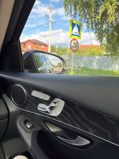 The image, taken from inside a car in Afanasovo, Russia, focuses on the interior door panel and the view through the window. The door panel features a speaker, intricate wood trim, and controls for seat adjustment and window operation. Through the window, a suburban landscape is visible, including a house with a red roof, a pedestrian crossing sign, a speed limit sign indicating 20 km/h, and lush green trees. The sky is blue with scattered clouds, and power lines are present in the background. The overall scene depicts a typical day in a residential area, viewed from the driver's or passenger's seat of a vehicle.