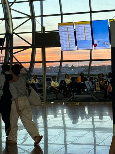 The image shows a group of people waiting in an airport terminal at sunset. The scene is viewed through a large, modern window with a grid of metal beams, revealing the warm hues of the sky outside. Several digital display screens are visible, showing departure information and flight details. People are seated on airport chairs, some looking at their phones, others conversing or resting. The overall atmosphere appears to be one of quiet anticipation. The lighting suggests it is late afternoon or early evening. The location is identified as Zaventem, Belgium.