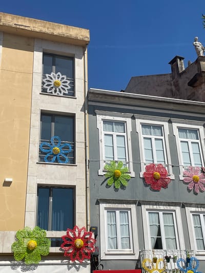 This media file showcases a vibrant urban scene in Águeda, Portugal, focusing on the colorful floral decorations adorning building facades. The image is dominated by the exteriors of two adjacent buildings. The building on the left, made of light yellow stone, features large windows framed by stone tiles. On the right, a gray building displays multiple floors of windows, each accented by large, intricate wire-frame flowers in bold colors such as pink, red, green, and blue. These decorative flowers, a signature element of Águeda's "AgitÁgueda" art festival, are suspended at various heights, adding a playful and artistic touch to the architecture. The sunlight suggests it is daytime, likely morning or afternoon, given the bright illumination and clear blue sky. The overall mood is cheerful and festive, reflecting the artistic atmosphere of the location. Visible architectural details include stone gargoyles on the upper left portion of the gray building, suggesting historical elements incorporated with modern decor. The scene appears to be a public street or plaza.