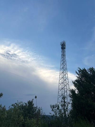 The image depicts a telecommunications tower situated in Wadern, Germany, against a backdrop of a cloudy sky. The tower, a tall, lattice-steel structure, is prominently positioned in the right half of the frame. It is equipped with numerous antennas at its apex. In the foreground, lush green foliage from trees and bushes partially obscures the base of the tower and the surrounding landscape. The sky features a mix of blue sky and wispy white clouds, with some darker cloud formations suggesting potential for changing weather. The overall scene is natural and serene, with no people or human activity visible.