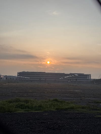 The image depicts a large, multi-story parking structure, likely part of an industrial complex or airport, set against a hazy, orange-tinted sky as the sun rises. The sun, a bright, almost perfectly round orb, is partially obscured by thin clouds. In the foreground, there is a flat, open area with sparse vegetation and what appears to be dark, gravelly soil. Several rows of solar panels are visible on poles in front of the parking structure, along with streetlights. The overall scene suggests an industrial or transportation hub in the early morning light, with a focus on infrastructure and natural elements. The location is identified as San Miguel Totoltepec, Mexico.