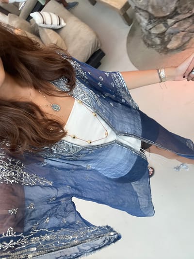 An overhead view captures an individual standing in a brightly lit lounge area within Psarou, Greece. The person wears a sheer, indigo-blue kaftan adorned with intricate silver beadwork and sequins, layered over a white top. Two distinct necklaces are present: one a gold-toned chain with a prominent oval-shaped pendant, and another a longer reddish cord featuring multiple gold spherical beads. A wide, textured gold-toned bracelet and a thinner cord bracelet are visible on the right wrist. A ring is worn on the left-hand ring finger. Dark sandals are partially visible on the feet. The immediate environment includes light-colored flooring, beige upholstered lounge seating with a striped cushion, and a textured, light-colored decorative column or natural element. A foot encased in a blue shoe is partially visible near the lounge seating in the background.