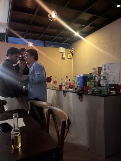 A low-angle indoor shot depicts a bar scene. Two individuals stand facing each other, one holding a smartphone, seemingly taking a photo of the other. A bar counter lines the wall behind them, stocked with liquor bottles, including Bacardi, and red plastic cups. A table with a wooden chair is to the right of the scene, where a beer bottle and white napkins are on it. Above, geometric light fixtures hang from the ceiling. The location and city of Santa Cruz Tehuixpango, Mexico, cannot be determined from the image.