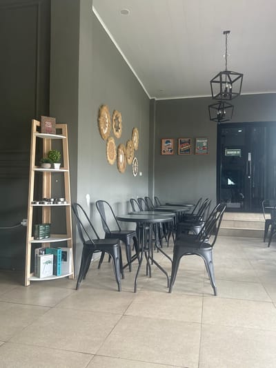 The image displays the interior of a modern cafe or coffee shop, devoid of people. A long row of black metal chairs and small, round tables is neatly arranged along the right side, with many chairs stacked or pushed under the tables, suggesting the establishment is closed or being prepared for service. The floor is covered with light-colored square tiles.
The walls are painted in two tones of grey; a darker shade forms the lower half, while a lighter grey, featuring horizontal paneling, covers the upper section on the left. Two geometric, black metal pendant lights hang from the ceiling. On the darker grey wall, a decorative arrangement of several circular woven mats with varying patterns and textures is visible. Above these, three framed pictures with vintage-style illustrations are displayed, one depicting a Vespa scooter and another with the text "KELLO".
In the foreground on the right, a tall, light-wood shelving unit holds various items including several books, a small potted plant, and other decorative objects. Visible book titles include "THE HEART OF COFFEE" and "MANIFESTO COFFEE", reinforcing the coffee-themed setting. At the back of the room, a glass door is visible, bearing the Indonesian text "RUANGAN AC" (Air-Conditioned Room), indicating the location is in an Indonesian-speaking region, consistent with the provided context of Sukabumi, Indonesia. The overall lighting suggests it is daytime, possibly outside operating hours.