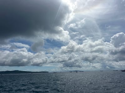 The image captures a wide view of the ocean and sky from the perspective of being on the water, likely from a boat, off the coast of Haad Yao, Thailand. The sea is a deep blue-gray, with small, white-capped waves reflecting the sunlight. The horizon is visible, with a lush, green, hilly island in the distance on the left, dotted with structures. Several longtail boats are scattered across the water, some appearing to be stationary. The sky is a dramatic mix of dark, heavy clouds and bright, sunlit patches. Streaks of sunlight break through the clouds, creating a dramatic lighting effect, with hints of a rainbow or atmospheric phenomenon near the brightest area of the sky. The overall scene suggests a dynamic weather day with intermittent sunshine and clouds.