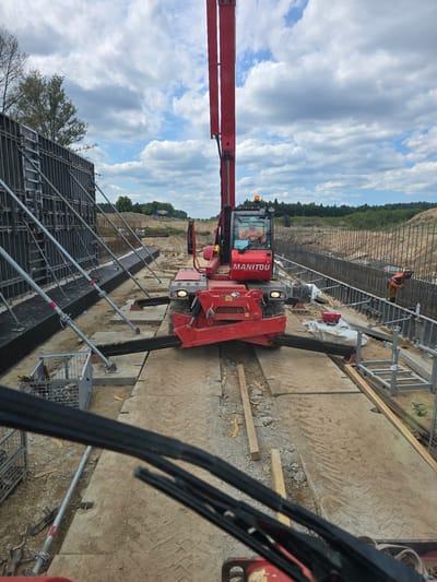 The media file depicts a construction site in Piecnik, Poland, under a partly cloudy sky. The scene features a red Manitou telehandler positioned on a dirt and gravel surface. The telehandler's boom is extended. In the background, a person wearing an orange high-visibility vest and a helmet is working near a structure reinforced with rebar and scaffolding. Another person, also in an orange vest, is visible inside the cab of the telehandler. The surrounding environment includes a tree line in the distance and more scaffolding and construction materials. The overall impression is of an active construction project. The word "MANITOU" is visible on the side of the telehandler.