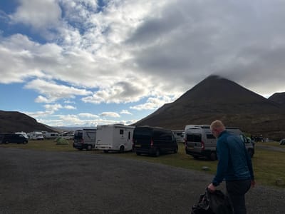 The image depicts a parking or camping area in the Isle of Skye, United Kingdom. Several camper vans and a few tents are scattered across a grassy area, adjacent to a gravel parking space. A mountain with a cloud-covered peak dominates the background. In the foreground, a man wearing a blue jacket and dark trousers is carrying a black bag. The sky is cloudy with patches of blue.