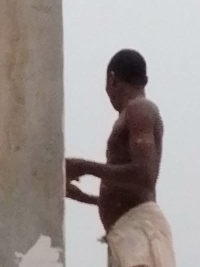 A shirtless dark-skinned man stands next to a large, gray, rectangular pillar. He is wearing white shorts. His head is turned away from the camera. The background is a pale, almost white color, suggesting an overcast day or a neutral backdrop. The man appears to be working or leaning against the pillar. His hands are positioned near the pillar. The image has a slightly blurry quality.
