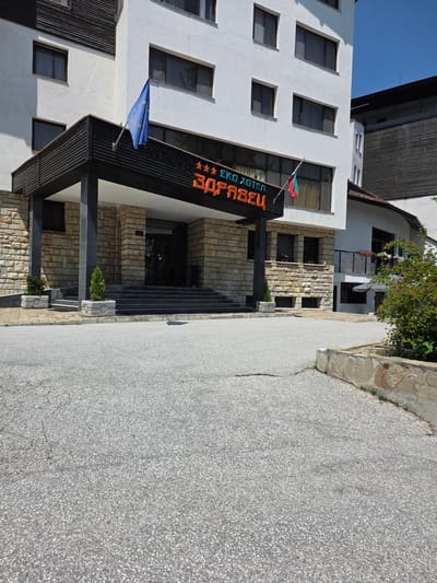 This is a medium shot, eye-level outdoor photograph of the entrance to the EKO Hotel Zdravets, located in Dedovo, Bulgaria. The hotel has a white facade with multiple windows. Above the entrance, a dark awning with the hotel's name in orange letters is visible, flanked by two flags: one the European Union flag and the other the Bulgarian flag. The entrance itself is accessed via a wide set of grey stairs leading to a double glass door. The building's base is constructed of stone blocks. Two small evergreen trees in grey planters are positioned on either side of the bottom of the stairs. A paved area, likely a parking lot or driveway, is in the foreground, with a low stone wall on the right side of the image. The sky is clear blue.