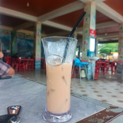 In Lhokdagang, Indonesia, a tall, clear glass filled with a light brown iced beverage, likely iced tea or coffee, sits on a textured grey surface. The glass is covered in condensation and contains several ice cubes. A black plastic straw extends from the top of the glass. A circular sticker on the glass reads "Crystal ONYX" with smaller text below indicating it is "Halal" and "Buatan Indonesia." In the foreground, to the left of the glass, a set of car keys with a BMW emblem is visible. The background is out of focus, showing the interior of an establishment with red chairs and tables. Pillars with colorful signage are also present. A person wearing a checkered shirt is partially visible on the left side of the frame. The environment suggests an outdoor or semi-outdoor seating area of a restaurant or cafe.