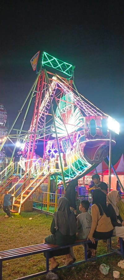 The image captures a night scene at a carnival in Banding Agung, Indonesia, dominated by a brightly lit Ferris wheel. The amusement ride is adorned with a vibrant array of neon lights in shades of green, pink, purple, and red, creating a festive and energetic atmosphere. The structure of the Ferris wheel is intricate, with a central support tower and spokes extending outwards, all outlined with glowing lights.
In the foreground, a group of people are seated on a long, blue bench, observing the carnival. Some are looking towards the Ferris wheel, while others are engaged in conversation. The individuals are dressed in casual attire, and several women are wearing hijabs. Their posture and expressions suggest a relaxed and enjoyable experience.
To the left of the Ferris wheel, a set of stairs leading up to the ride is also illuminated with colorful lights, suggesting that the carnival is in full swing. In the background, hints of other carnival structures and tents are visible, further establishing the setting as a lively amusement park. The ground is covered with grass, and the overall scene is illuminated by the artificial lights of the carnival, casting a warm glow.
The time of day is clearly nighttime, indicated by the dark sky and the prominence of artificial lighting. The weather appears to be clear, as there are no signs of rain or adverse conditions. The image evokes a sense of community and shared enjoyment, as people gather to partake in the festivities of the carnival. There is no visible text in the image.
