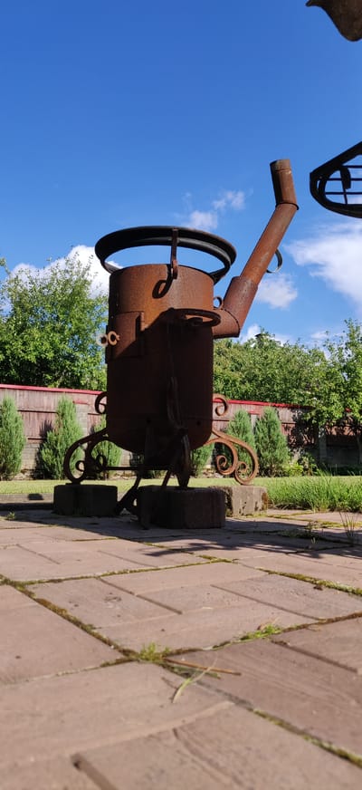 This is a low-angle, medium shot of a rusty, ornate metal sculpture, possibly a repurposed industrial object or a decorative garden feature, in an outdoor setting. The sculpture, which appears to be made of thick metal sheets, has a cylindrical body with a wide, open top rim. It is supported by decorative metal scrollwork legs that terminate in wheels. A pipe extends upwards and to the right from the main body, resembling a spout or chimney. There are two circular openings on the front of the cylinder. The sculpture is placed on a paved area made of reddish-brown bricks, with some grass and weeds growing between the stones. Behind the sculpture, a wooden fence with a red top runs horizontally, and beyond that, there are lush green trees and bushes. The sky above is a bright blue with scattered white clouds. The overall impression is one of a sunny day in a garden or yard. The location is identified as Калодзішчанскі сельскі Савет, Belarus. No people are present in the image, and no specific actions or interactions are depicted.