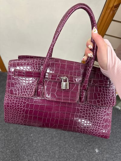 A person's hand with white nail polish and floral designs is holding a shiny, plum-colored handbag with a crocodile-like texture. The handbag has a silver lock on the front flap and two top handles. The background is a plain white wall with a wooden beam visible to the right. The handbag is positioned in the foreground, obscuring most of the person's arm. The image does not provide any specific visual cues to indicate a location in Carlow, Ireland.