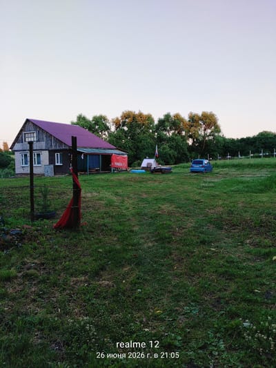An exterior, eye-level, wide shot captures a rural landscape at dusk, with a wooden house and a blue car as the main subjects. The house, positioned on the left, is a two-story structure with a distinctive purple roof. Its exterior walls appear to be a combination of brick and wood. In front of the house, a wooden fence post stands, adorned with a red cloth. To the right of the house, a blue hatchback car is parked in a grassy field. Further to the right, a small white tent is set up, with a Russian flag planted next to it. The environment is characterized by a vast expanse of green grass, with a line of trees forming a backdrop behind the house and car. The sky is a pale blue, suggesting the transition from day to night. A timestamp in the lower right corner indicates "realme 12 26 June 2026 at 21:05," providing a temporal context for the scene.