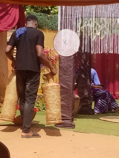 In Ochadam, Nigeria, a man stands in an outdoor, decorated setting, viewed from the back. He wears a black short-sleeved t-shirt, dark trousers, and dark sandals, with a blue patterned cloth draped over his left shoulder. He holds a tall, cylindrical, light-brown woven container in each hand. To his right, partially visible, a person is seated on the ground, wearing a blue top and a purple patterned wrapper. The background features red drapes on the left and right, framing a dark vertical fabric column from which multiple white strings hang. A circular, light-colored woven disc with a spiral pattern is affixed to this dark column. Behind the man, a yellow wall is partially visible, adorned with colorful artificial flowers. Green foliage is present further in the background. The ground consists of bare earth in the foreground and a green mat extending towards the right.