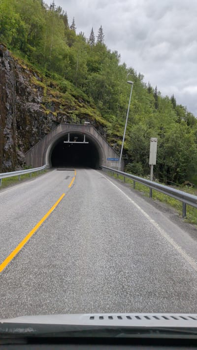 This media file shows a view from a car driving towards the Hjartåsen Tunnel in Mosjøen, Norway. The tunnel entrance is a large, arched concrete structure carved into a rocky, tree-covered mountainside. The road leading to the tunnel is paved and has a yellow double line marking the center. There are metal guardrails along the sides of the road. A tall, modern lamppost stands to the right of the road, and a vertical white information box with several panels is mounted on a pole nearby. Above the tunnel entrance, a dark-colored structure with lights is visible. The sky is overcast, suggesting a cloudy day. The surrounding landscape is lush with green trees and vegetation, typical of a mountainous, possibly forested, Norwegian environment. The overall impression is of a journey through a natural landscape towards a man-made infrastructure. There is a blue sign to the right of the tunnel entrance that reads "Hjartåsen Tunnel". The bottom of the image shows the dashboard of the car. No people are visible.