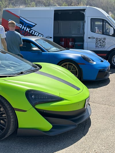 The image shows a white van parked next to a blue sports car and a lime green sports car. The van has a logo of "TITANKLAV RACING" and a QR code. A man in a grey shirt is standing near the van and the blue car. The cars are parked on an asphalt surface. The location, Sadovoe, Russia, is not verifiable in the image.