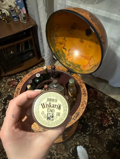 The image captures a top-down view of a person's right hand holding a small bottle, positioned above an open globe-shaped bar. The scene is set indoors, likely a home in Krapivka, Russia, with a dark, patterned rug covering the floor.
The main subjects include the hand and the bottle, an antique-style globe bar, and various miniature bottles. The globe bar is open, with its top half resting beside the base. The interior of the globe's top half features an aged map or artistic depiction with classical figures. The circular base of the globe bar serves as a rack, holding several small liquor bottles upright.
The bottle held by the hand has a white label with prominent black Cyrillic text that reads "Wiskarik." Below this, "40%" is clearly visible, indicating the alcohol content, and "0.1л" denotes a volume of 0.1 liters. Other smaller Cyrillic text, a QR code, a barcode (4606034507576), and an "EAC" (Eurasian Conformity) mark are also visible on the label. The hand displays light-colored nail polish and a small, faded tattoo on the side of a finger.
To the left of the globe bar, on a light-colored surface, several other small, clear glass bottles, some with blue labels, are arranged. Further left and partially obscured, a wooden cabinet or shelf unit containing books or other items is visible. The carpet underneath features intricate floral and geometric patterns in shades of red, white, and dark blue/black.
The lighting in the image is dim, suggesting it was taken during the evening or night under artificial light. No specific activity beyond holding the bottle and its proximity to the globe bar is evident.