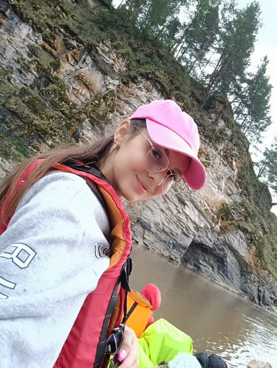 A young woman with long brown hair, wearing a pink baseball cap and round glasses, is taking a selfie from a raft on a river. She is wearing a grey sweatshirt and a red and orange life vest. The background shows a steep, rocky cliff face covered in moss and trees, with the water of the river in the foreground. The image appears to have been taken in Idrisovo, Russia, based on the provided information.