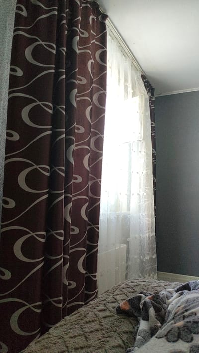 The image depicts a bedroom interior, likely located in Monino, Russia. On the left side of the frame, a dark brown curtain with a white swirling pattern is visible, partially obscuring a window. The window is covered with a sheer white curtain with a delicate floral pattern, allowing diffused sunlight to enter the room. To the right of the window, a dark gray wall forms the background. In the foreground, a bed is covered with a textured, light brown blanket and a patterned throw blanket. The overall impression is a dimly lit, domestic space, possibly a bedroom. There are no visible people or discernible actions. The lighting suggests it is daytime, with natural light filtering through the window.