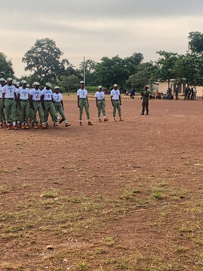 On an open, reddish-brown dirt field with sparse green patches in Kubi A, Nigeria, approximately 17-20 individuals are present. Fourteen individuals are observed in the foreground, arranged in a formation or line. They wear white t-shirts with "NYSC" branding, green trousers, and brown boots, with some also wearing matching green caps. One individual in this group has their left leg extended forward, mid-action. To their right and slightly forward, a single individual in a full military camouflage uniform and a red beret stands facing the formation, appearing to supervise or instruct. In the mid-ground and background, numerous trees are visible, along with utility poles and indistinct buildings. To the far right in the background, another group of people is present, some appearing to be seated or standing under trees. The sky is overcast and light gray.